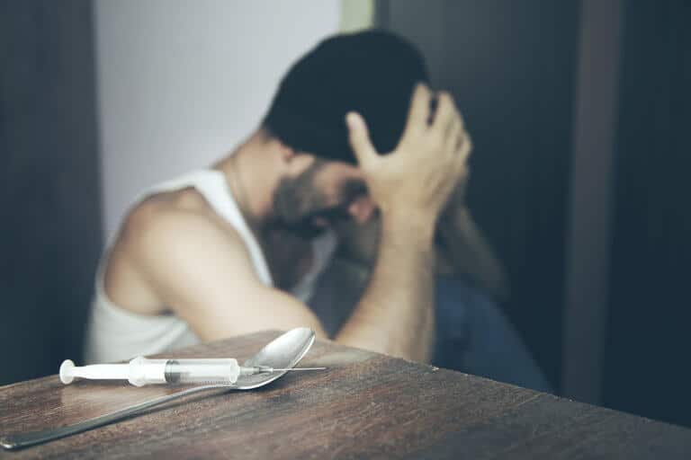 man in beanie on floor with his hands on his head and needle with spoon on table as man tries to quit heroin cold turkey